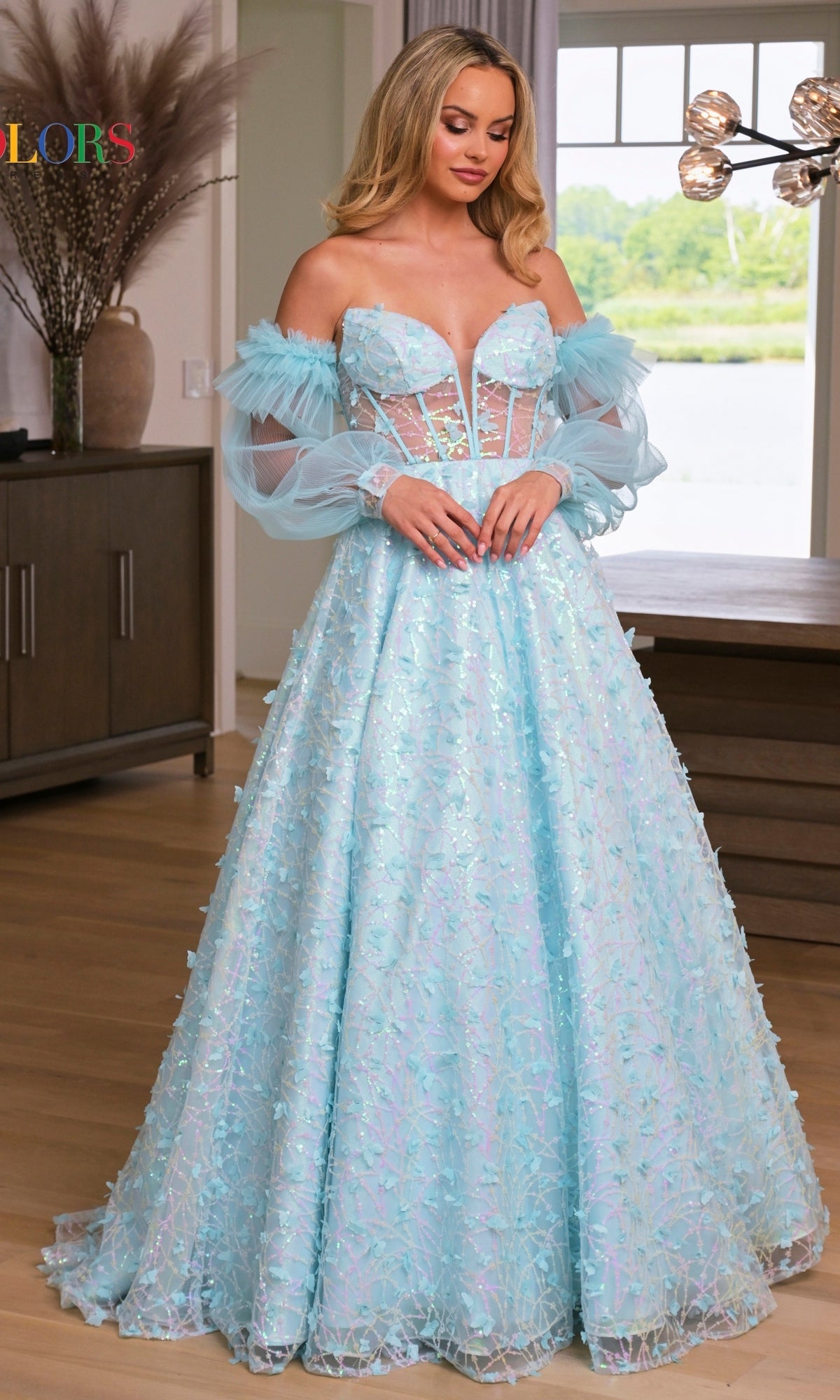 A woman stands indoors wearing the 3D Butterfly Formal Ball Gown 3226, a light blue, floor-length ball gown with a strapless corset-style bodice and removable sheer sleeves.